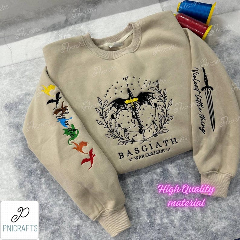 K&ouml;nnte beinhalten: Ein beiger Pullover mit einem schwarz-wei&szlig;en Motiv eines Drachen und eines Schwertes auf der Vorderseite. Der Text "BASGIATH" und "WAR COLLEGE" ist unter dem Motiv gedruckt. Der linke &Auml;rmel hat eine Reihe von bunten gestickten Drachen.