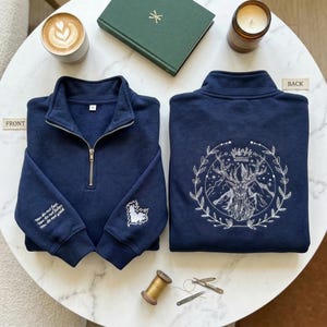 Könnte beinhalten: Marineblaues Sweatshirt mit Viertelreißverschluss und weißen Stickereien. Auf der Vorderseite befinden sich eine Karte und Text, auf der Rückseite ein Hirsch mit Krone und Berglandschaft. Kaffee, Buch und Kerze sind ebenfalls zu sehen.