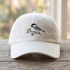 Peut inclure: Casquette de baseball blanche avec un motif brodé d'un petit oiseau perché sur une branche avec des feuilles vertes. L'oiseau a des plumes noires, blanches et grises. La casquette est en tissu doux et a une visière incurvée.
