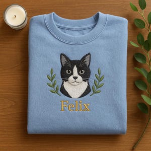 May include: A light blue sweatshirt featuring an embroidered black and white cat face with the name "Felix" in gold lettering. The cat is surrounded by a green olive branch design. The sweatshirt is folded on a wooden surface.
