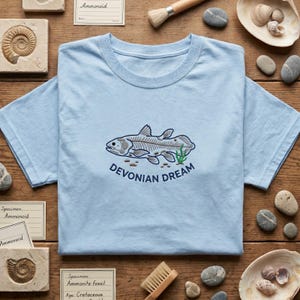 May include: A light blue t-shirt featuring an embroidered graphic of a prehistoric fish skeleton with the text DEVONIAN DREAM in dark blue lettering below the fish, accompanied by small brown pebbles and a green aquatic plant illustration.