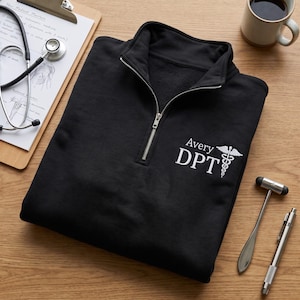 May include: A black quarter-zip pullover featuring white embroidered text that reads Avery DPT alongside a caduceus symbol on the left chest. The garment has a high collar with a silver-toned metal zipper closure and long sleeves.