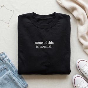 May include: Black crew neck sweatshirt with the white text "none of this is normal". The image shows the sweatshirt with blue jeans, a silver necklace, and white trainers.