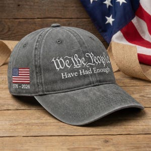 May include: A charcoal grey baseball cap featuring the white embroidered text We The People Have Had Enough. The side displays a small embroidered American flag with the white text 1776-2026 positioned directly underneath the flag graphic.