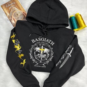 Op de afbeelding: Zwart hoodie met gouden borduurwerk op de mouwen. De voorkant van de hoodie heeft een witte afbeelding met de tekst "BASGIATH WAR COLLEGE DRAGON RIDER".