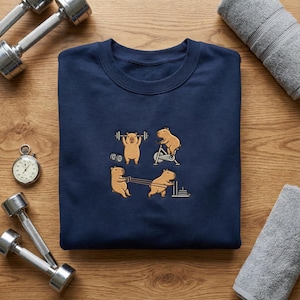 May include: Navy blue sweatshirt featuring a graphic of capybaras working out with weights, on a stationary bike, and doing other exercises. The sweatshirt is surrounded by dumbbells, a stopwatch, and a gray towel.