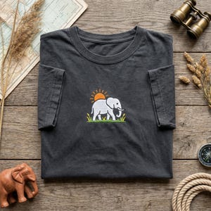 Peut inclure: Un t-shirt gris anthracite orné d'un motif brodé central représentant un éléphant blanc marchant dans de l'herbe verte sous un soleil orange. Le t-shirt présente un col rond et des manches courtes, avec un design sobre et épuré.