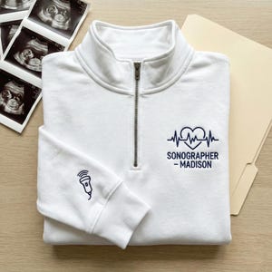 May include: A white quarter-zip sweatshirt with navy blue embroidery. The design features a heart-shaped EKG, a microphone, and the text "SONOGRAPHER - MADISON". The sweatshirt is folded on a wooden surface, with sonogram images and a file folder.