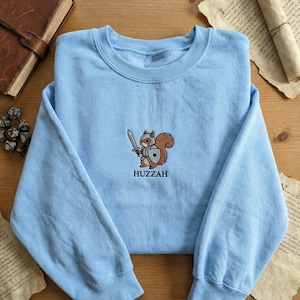 May include: Light blue sweatshirt with an embroidered design of a squirrel in armor holding a sword and shield. The word "Huzzah" is embroidered below the squirrel. The sweatshirt is made of a soft material.