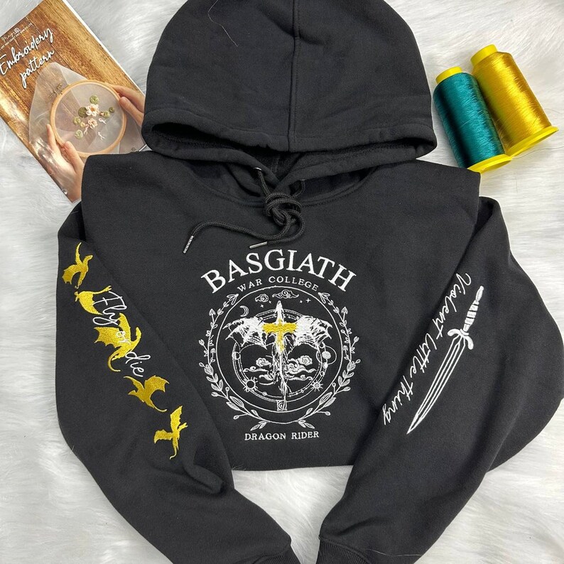 Op de afbeelding: Zwart hoodie met een wit grafisch ontwerp aan de voorkant met de tekst "Basgiath War College Dragon Rider" met een zwaard en een draak afbeelding. De mouwen zijn geborduurd met een draak en een zwaard in goud.