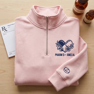 Peut inclure: Un sweat-shirt rose clair à fermeture éclair quart de zip avec un motif de pilule bleu marine et blanc en forme de cœur, avec le texte "Pharm D-Amelia". Un carnet de prescriptions et des flacons de pharmacie sont en arrière-plan.