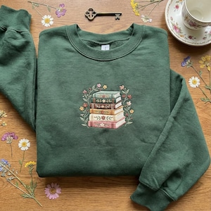 May include: A dark green sweatshirt featuring an embroidered design of a stack of books surrounded by a floral wreath. The books have different colors and floral details. The sweatshirt is on a wooden surface.