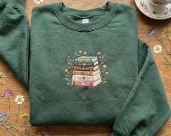 May include: A dark green sweatshirt featuring an embroidered design of a stack of books surrounded by a floral wreath. The books have different colors and floral details. The sweatshirt is on a wooden surface.