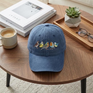 May include: A blue denim baseball cap with an embroidered design of five colorful birds. The birds are in shades of brown, yellow, green, orange, and blue. The cap is sitting on a dark brown wooden table.