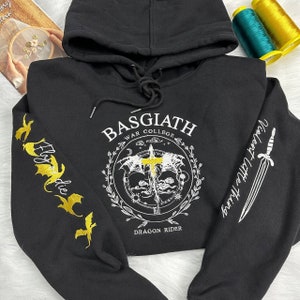 Op de afbeelding: Zwarte hoodie met een wit geborduurd ontwerp aan de voorkant met de tekst "Basgiath War College Dragon Rider". De linkermouw heeft een gouden geborduurd drakenontwerp en de rechtermouw heeft een wit geborduurd zwaardontwerp.