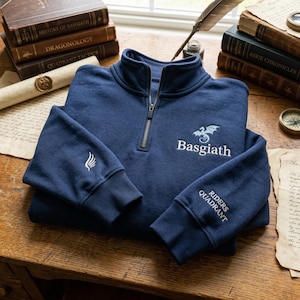 May include: A navy blue quarter-zip sweatshirt with embroidered text and a dragon design. The sleeve features "Riders Quadrant" and a wing emblem. The sweatshirt is displayed on a wooden surface with books and parchment.