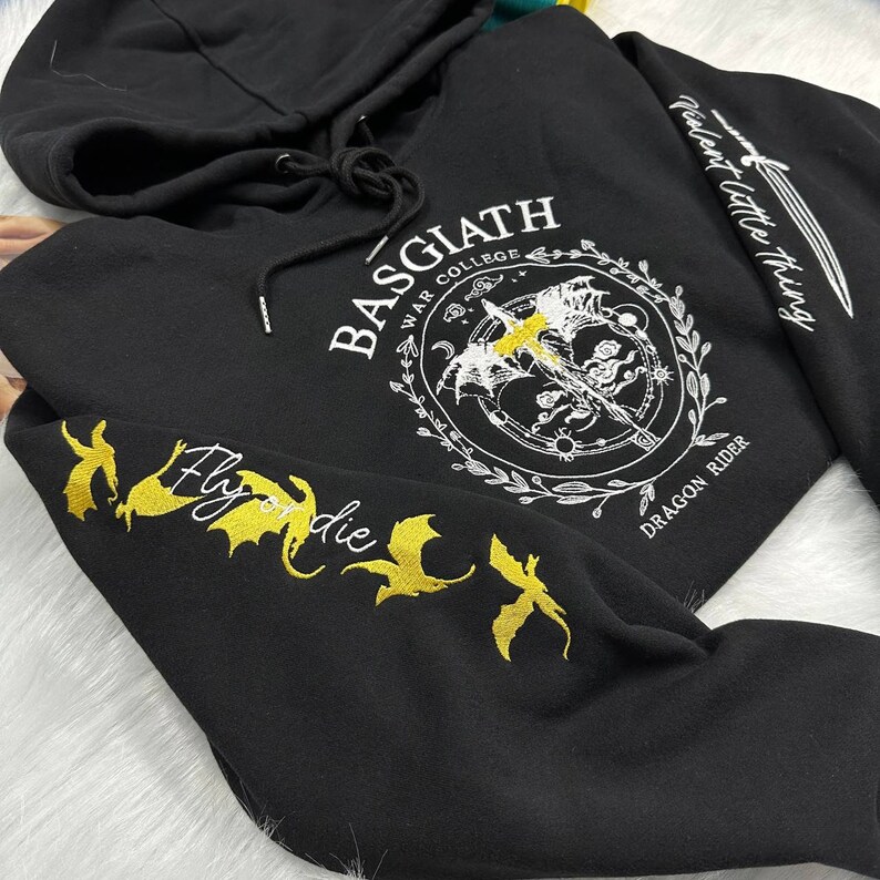 Op de afbeelding: Zwarte hoodie met een wit geborduurd ontwerp aan de voorkant dat "Basgiath War College Dragon Rider" zegt en een wit geborduurd ontwerp op de rechtermouw dat "Violent Little Thing" zegt. De linkermouw heeft een gouden geborduurd ontwerp van draken en de woorden "Fly or die".