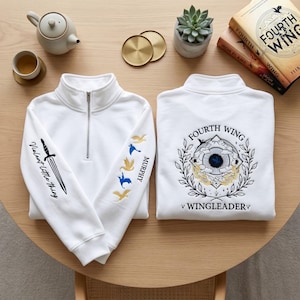 K&ouml;nnte beinhalten: Wei&szlig;es Sweatshirt mit Viertelrei&szlig;verschluss und gestickten Details. Der linke &Auml;rmel zeigt ein Schwert und Text, w&auml;hrend der rechte &Auml;rmel Vogelgrafiken und den Namen "Murphy" aufweist. Auf der R&uuml;ckseite befindet sich ein kreisf&ouml;rmiges Design mit dem Text "Fourth Wing" und "Wingleader".
