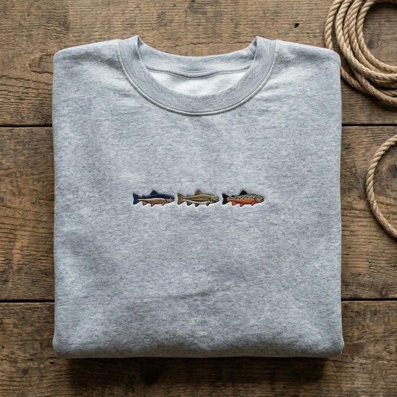 May include: A light gray sweatshirt featuring three embroidered fish in blue, green, and orange. The sweatshirt is folded neatly on a wooden surface. The design is simple and nature-themed.