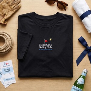 May include: Black t-shirt with the text "Monte Carlo Sailing Club" in white and blue. The design includes a red flag, a yellow sun, and blue waves. The shirt is surrounded by nautical accessories, including a rope, sunglasses, and a bottle of sparkling water.