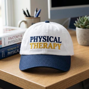 May include: A white and navy blue baseball cap with the words "PHYSICAL THERAPY" embroidered in navy blue and yellow. The cap is sitting on a wooden desk next to books and a small potted plant.