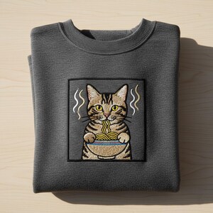 May include: A dark gray sweatshirt with a square embroidered patch featuring a cartoon cat eating noodles from a blue bowl. The cat is brown and tan with yellow eyes. Steam rises from the bowl. The sweatshirt is folded.