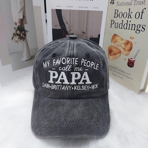 May include: A charcoal gray baseball cap with a distressed finish featuring white embroidered text that reads MY FAVORITE PEOPLE call me PAPA followed by the names DAIN, BRITTANY, KELSEY, and NICK in a horizontal line across the front panel.