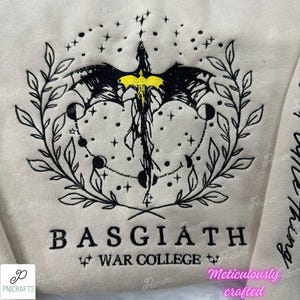K&ouml;nnte beinhalten: Ein beiger Pullover mit einem schwarzen gestickten Design eines Drachen mit gelben Akzenten, umgeben von einem Kranz aus Bl&auml;ttern und Sternen. Der Text "BASGIATH WAR COLLEGE" ist unter dem Design gestickt.