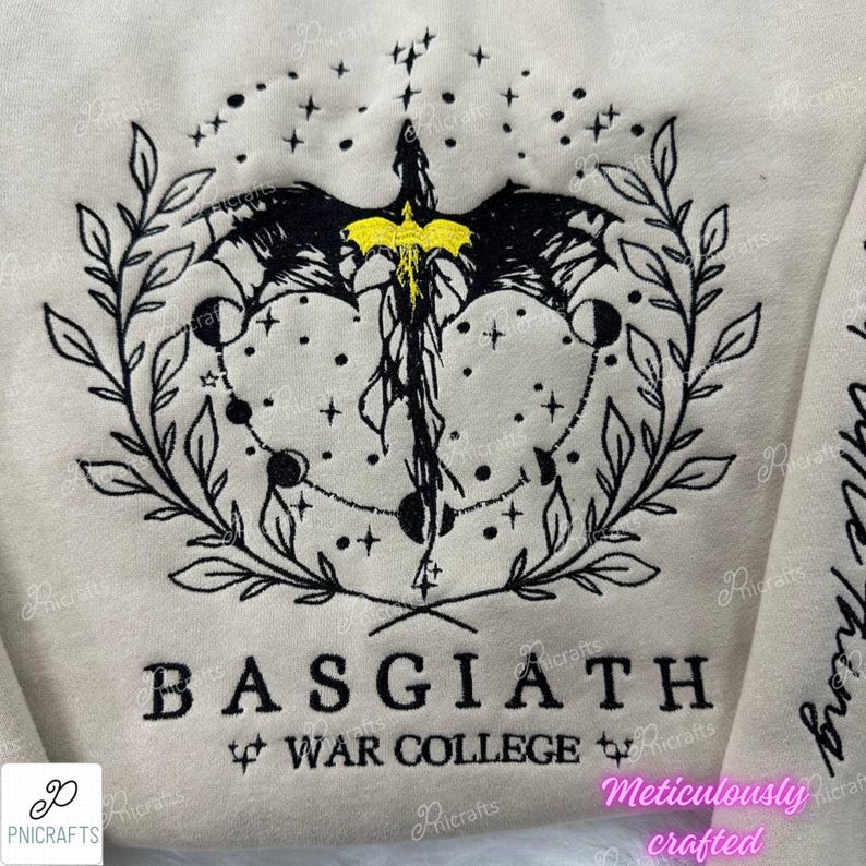 K&ouml;nnte beinhalten: Ein beiger Pullover mit einem schwarzen gestickten Design eines Drachen mit gelben Akzenten, umgeben von einem Kranz aus Bl&auml;ttern und Sternen. Der Text "BASGIATH WAR COLLEGE" ist unter dem Design gestickt.