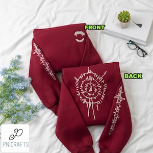 May include: Maroon sweatshirt with white embroidered designs. The front features a toothy grin, while the back displays a circular pattern. Sleeves have linear and cross designs. The text "FRONT" and "BACK" are visible.