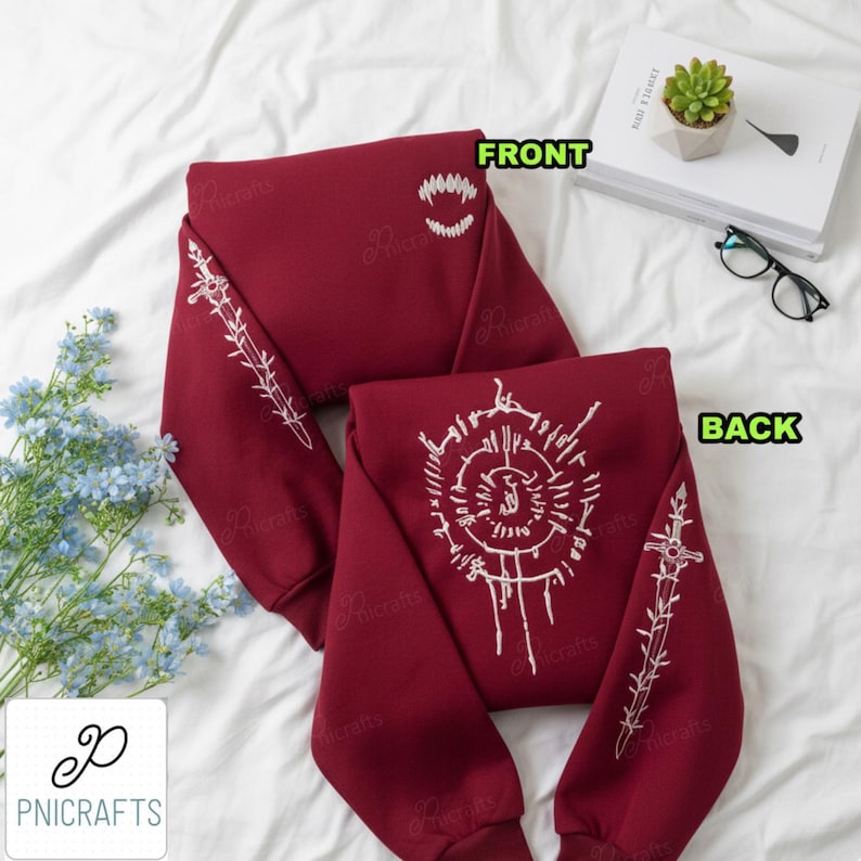 May include: Maroon sweatshirt with white embroidered designs. The front features a toothy grin, while the back displays a circular pattern. Sleeves have linear and cross designs. The text "FRONT" and "BACK" are visible.