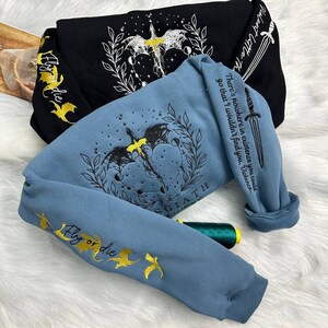 May include: Two sweatshirts with embroidered designs. One is black with a white dragon and the other is blue with a black dragon. Both have the text "Fly or die" embroidered in gold on the sleeves. The blue sweatshirt also has the text "There's nowhere in existence you could go that I wouldn't find you, Violence" and "SGIATH" embroidered on the front.