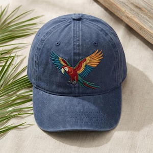 Könnte beinhalten: Eine blaue Baseballkappe mit einem gestickten Papageien-Design. Der Papagei hat rote, gelbe und blaue Flügel. Die Kappe ist aus weichem, gewaschenem Baumwollmaterial.