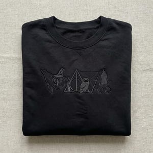 May include: Black sweatshirt with embossed Harry Potter-themed imagery. Includes a broom, sorting hat, Deathly Hallows symbol, owl, candle, wizard, and glasses. The number 9 3/4 is also visible. The sweatshirt is neatly folded.