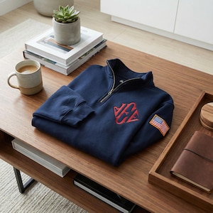May include: Navy blue quarter-zip sweatshirt with a red embroidered logo and an American flag patch on the sleeve. The sweatshirt is folded on a wooden table, alongside a mug, books, and a small succulent plant.