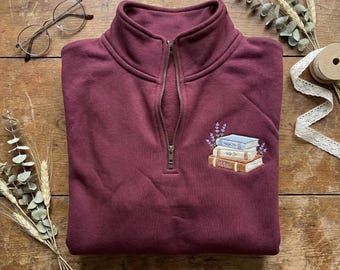 May include: A burgundy quarter-zip sweatshirt with an embroidered design of stacked books and lavender sprigs. The sweatshirt has a high collar and a silver zipper. A pair of round eyeglasses and decorative sprigs are on the wooden surface.