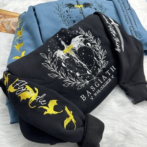 May include: Black sweatshirt with a white graphic of a dragon with wings spread, surrounded by a wreath and moons. The text "BASGIATH WAR COLLEGE *+*" is printed below the dragon. The sweatshirt has gold embroidered dragons on the sleeves with the text "Fly or die".