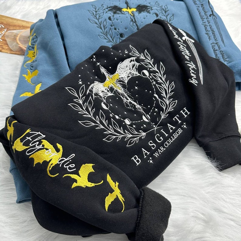 May include: Black sweatshirt with a white graphic of a dragon with wings spread, surrounded by a wreath and moons. The text "BASGIATH WAR COLLEGE *+*" is printed below the dragon. The sweatshirt has gold embroidered dragons on the sleeves with the text "Fly or die".