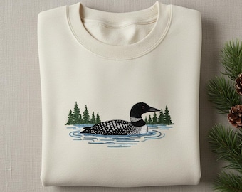 May include: Cream-colored sweatshirt featuring an embroidered design of a loon swimming in a lake. The loon has black and white markings, with green trees in the background. The design is centered on the front of the folded sweatshirt.