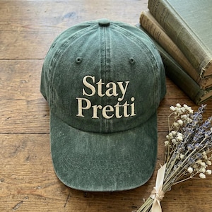 May include: A green baseball cap with the embroidered text "Stay Pretti" in white. The cap is made of a soft, textured fabric. The background includes vintage books and dried flowers.