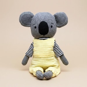 Puede incluir: Una muñeca de koala de peluche hecha a mano. El koala tiene pelaje gris, orejas grandes y redondas y una nariz negra. Lleva un mono amarillo sobre una camiseta a rayas blancas y negras. La muñeca está sentada sobre un fondo neutro.