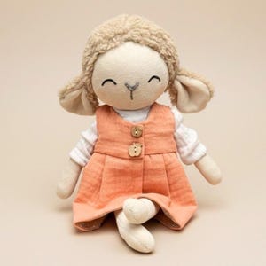 Puede incluir: Una muñeca de peluche de cordero con cabeza y orejas texturizadas de color marrón claro, vestida con un vestido naranja con botones de madera sobre una camisa blanca. La muñeca tiene una cara sonriente y las piernas cruzadas.