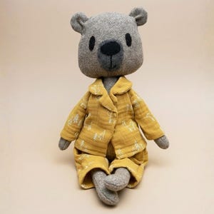 Puede incluir: Un muñeco de oso de peluche con cabeza y cuerpo grises, vestido con pijama amarillo con un estampado de árboles blancos. El oso está sentado con las piernas cruzadas. El fondo es de color beige liso.