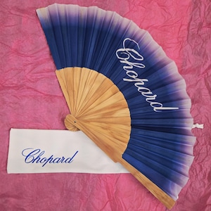 May include: A folding hand fan with a light wood frame and a fabric leaf that features a blue to white ombre gradient. The word Chopard is printed in white script on the fan and in blue script on an accompanying white rectangular fabric sleeve.