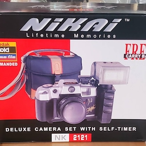 Op de afbeelding: Een zwarte en zilveren 35mm filmcamera met een ingebouwde flitser en een zwarte draagtas. De doos heeft de tekst "Nikai Lifetime Memories" en "Deluxe Camera Set with Self-Timer NK 2121".