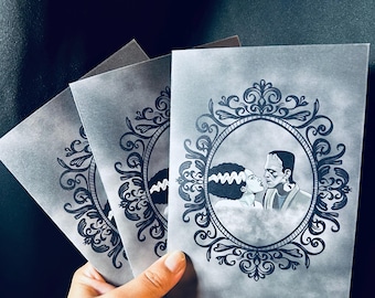 May include: Three gray cards featuring a black and white illustration of the Bride of Frankenstein and Frankenstein's monster in an ornate oval frame. The cards are held in a hand against a dark background.