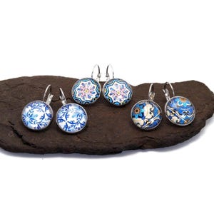 May include: Three pairs of round silver-tone leverback earrings featuring glass cabochons with intricate blue, white, and pink floral and geometric patterns. Each earring displays a distinct decorative tile-inspired design in a circular setting.