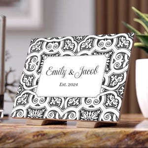 Op de afbeelding: Een decoratieve houten fotolijst met een zwart-wit geometrisch patroon. De lijst bevat de namen "Emily & Jacob" en het jaar "Est. 2024" in elegant schrift. De lijst wordt ondersteund door twee kleine houten standaards.