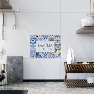 May include: A decorative tile arrangement with a white rectangular center displaying the text "FAM&Iacute;LIA ROCHA" in a blue and yellow floral pattern. The tiles are set against a white wall in a kitchen setting.