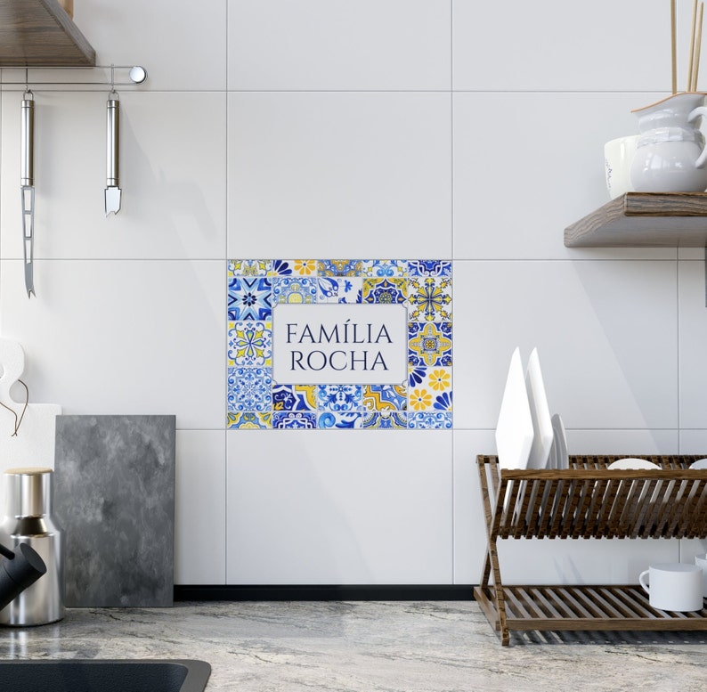 May include: A decorative tile arrangement with a white rectangular center displaying the text "FAM&Iacute;LIA ROCHA" in a blue and yellow floral pattern. The tiles are set against a white wall in a kitchen setting.